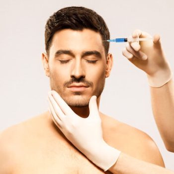 Young shirtless man receiving beauty injection in forehead to remove wrinkles, face held by hands of cosmetologist, isolated on gray background. Plastic surgery concept